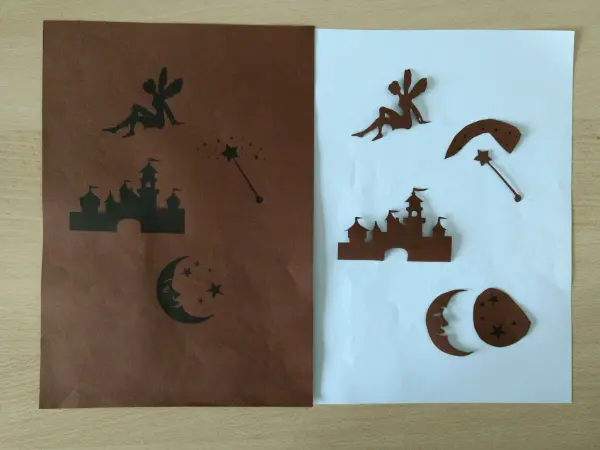 Schwarze Bastelschnitte von Feen, Mond und Burgen liegen auf braunem und weißem Papier, bereit für das Windlicht „Fee im Glas“.