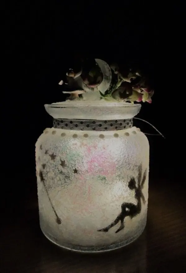 Ein transparentes Windlicht mit einer Fee und Sternenmotiven leuchtet sanft im Dunkeln und ist mit Blumen dekoriert.