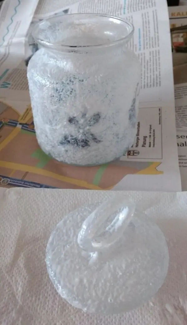Ein gläsernes Windlicht mit einer transparenten, glitzernden Oberfläche steht auf einem Tisch, umgeben von Zeitungspapier und Bastelmaterial.