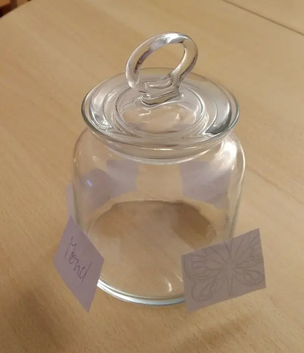 Ein rundes, transparentes Glas mit einem Deckel steht auf einem Tisch und wartet darauf, als Windlicht „Fee im Glas“ dekoriert zu werden.