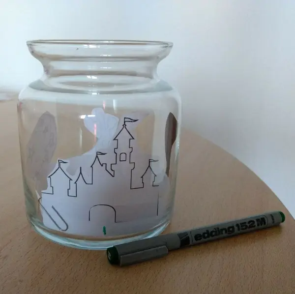 Ein transparentes Windlicht zeigt eine skizzierte Burg im Inneren. Daneben liegt ein Stift zum Ausmalen auf einem Tisch.
