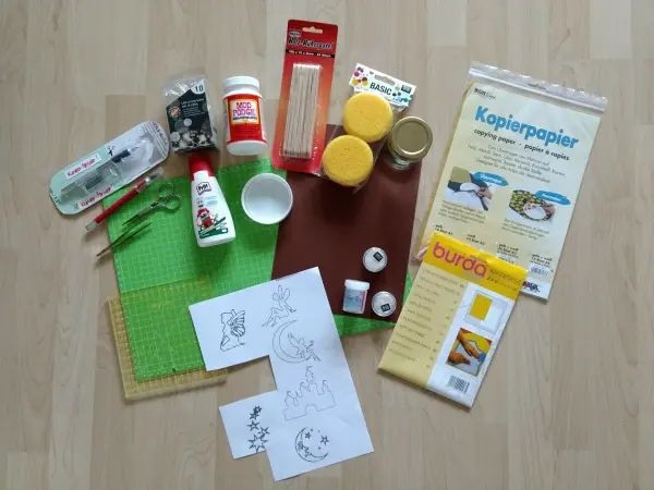 Bastelmaterialien für ein Windlicht „Fee im Glas“ liegen auf einem Tisch. Dazu gehören Papiere, Glasgefäße, und Bastelkleber.