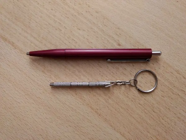 Ein roter Kugelschreiber liegt neben einem silbernen Schraubendreher für Brillen, der an einem Schlüsselring befestigt ist.