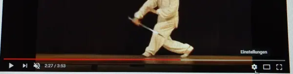 Ein Tänzer in beiger Kleidung führt eine elegante Bewegung auf einer Bühne aus, während ein Video abgespielt wird.