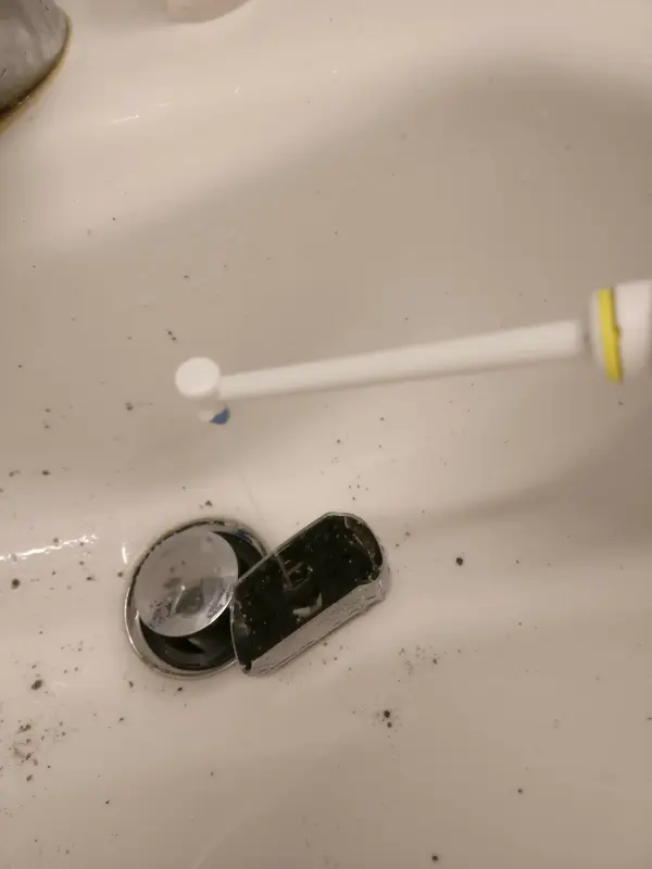 Eine Hand hält eine Zahnbürste, während sie Reinigungskartuschen für Rasierer in einem Waschbecken säubert. Unter der Zahnbürste liegt Schmutz.