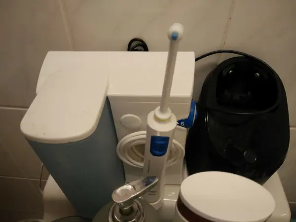 Ein Rasierer mit Reinigungskartuschen steht auf einem Badewaschbecken neben einer elektrischen Zahnbürste und einem schwarzen Gerät.