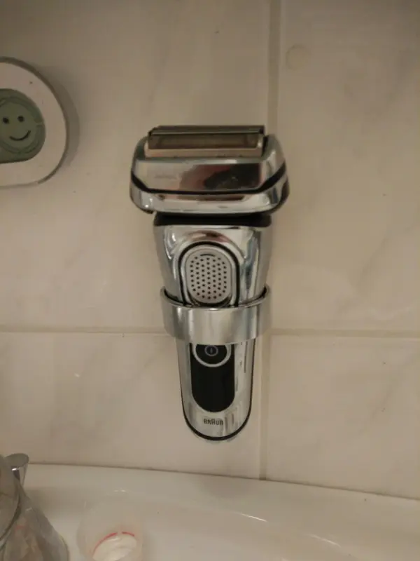 Ein Rasierer hängt an der Wandhalterung, bereit zur Nutzung im Badezimmer, mit Fliesen im Hintergrund.