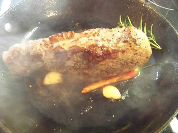 Ein saftiges Roastbeef vom Limousin Rind brät in einer Pfanne mit Kräutern und Knoblauch, umgeben von aufsteigendem Dampf.