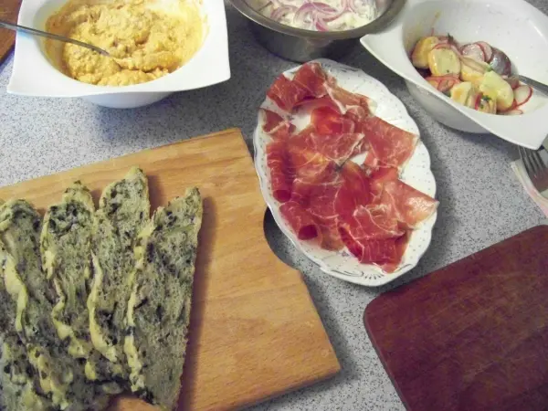 Ein dunkles Brot mit Mangold liegt auf einem Holzbrett, umgeben von verschiedenen Dips und Schinken auf einem Tisch.