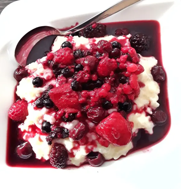 Cremiger Milchreis ist in einer weißen Schale mit einer Mischung aus roten Beeren und Sauce angerichtet, dekoriert mit einem Löffel.