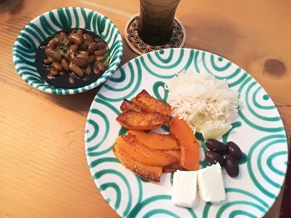 Ein Teller mit gebratenem Kürbis, Reis und Feta liegt neben einer Schüssel mit getrocknetem Bohnensalat auf einem Holztisch.