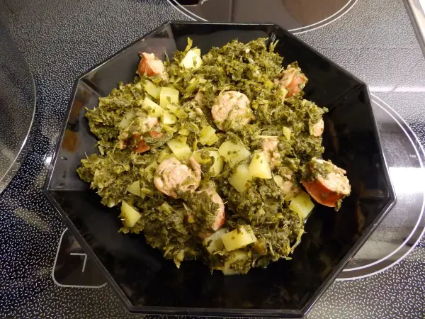 Eine Schüssel Grünkohl mit Mettwurst und Kartoffeln steht auf einem platten Herd, gut angerichtet und dampfend heiß.