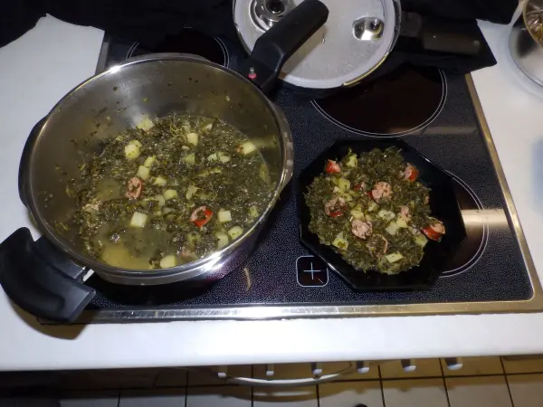 Ein großer Topf mit Grünkohl, Kartoffeln und Mettwurst kocht auf dem Herd, während eine kleine Pfanne daneben gefüllt ist.