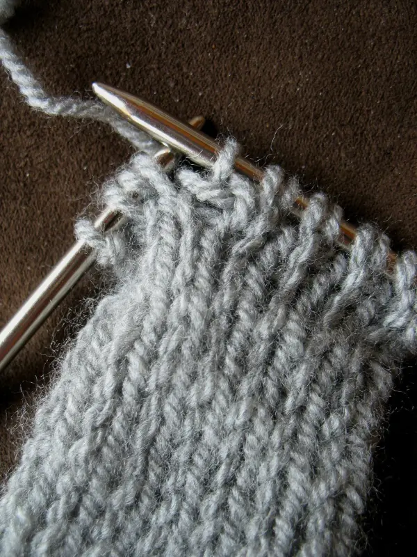 Ein graues selbst gestricktes Stirnband wird mit silbernen Stricknadeln gearbeitet, auf braunem Stoff liegend.
