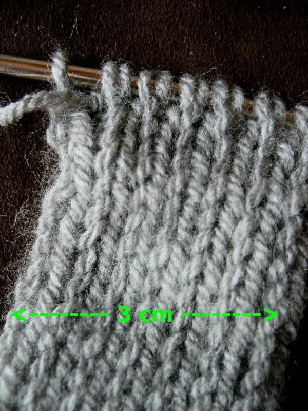 Ein graues, selbst gestricktes Stirnband liegt auf einer dunklen Oberfläche, während Wolle und eine Stricknadel sichtbar sind.