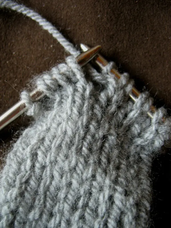 Ein graues, selbst gestricktes Stirnband wird auf einer Stricknadel gefertigt, umgeben von einem braunen Untergrund.