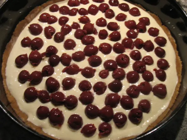 Eine Quark-Sahne-Kirsch-Torte mit Kokosgeschmack steht auf einem schwarzen Kuchenblech, bedeckt mit vielen Kirschen.