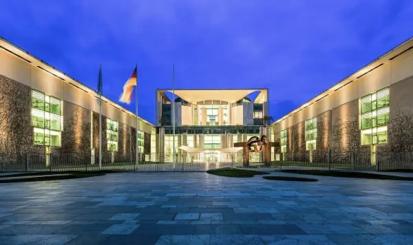 Ein modernes Gebäude mit Glasfassade steht im Zentrum, umgeben von einem klaren Abendhimmel und einer gepflasterten Fläche.