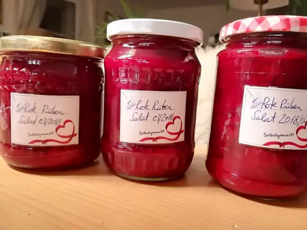 Drei Gläser mit roten Rüben im Glas, frisch konserviert und auf einem Holzstamm platziert. Labels beschreiben den Inhalt.