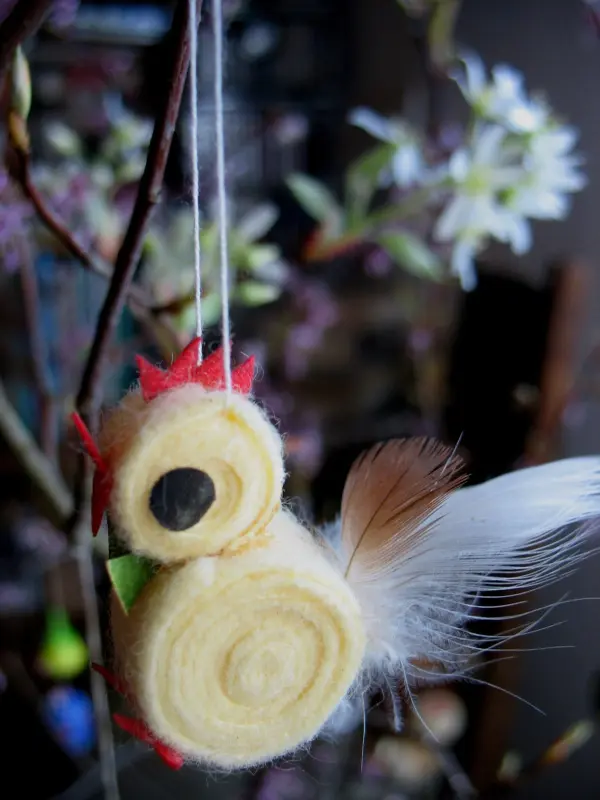 Ein buntes, handgefertigtes Hühnchen baumelt an einem Zweig, umgeben von blühenden Ästen und Frühlingsdeko.
