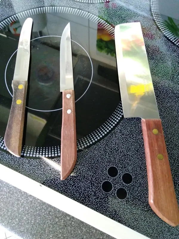 Drei scharfe Messer mit Holzgriffen liegen auf einer schwarzen Herdplatte, bereit für das Schneiden von Zutaten.