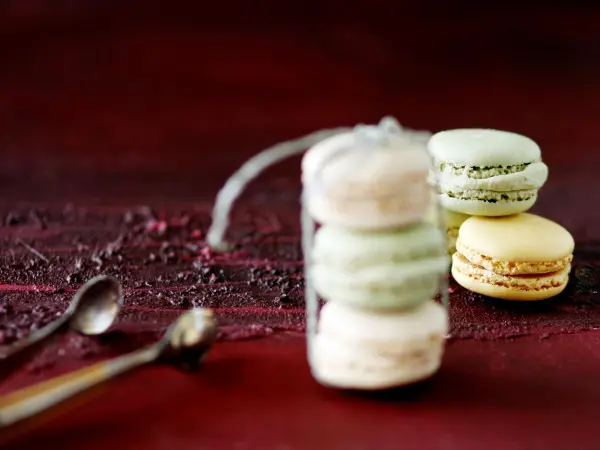 Zwei farbige Macarons stehen auf einem dunklen Untergrund, während der Fokus auf einem Glasbehälter mit weiteren Macarons liegt.