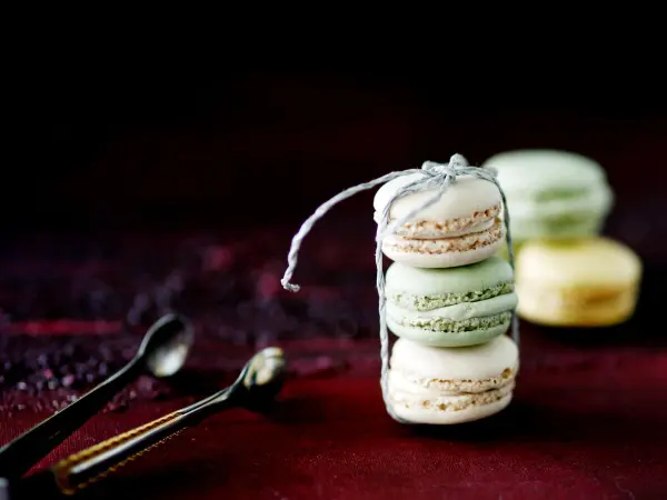 Drei farbige Macarons stehen in einer transparenten Verpackung und sind von silbernen Löffeln umgeben, auf einem dunklen Untergrund.