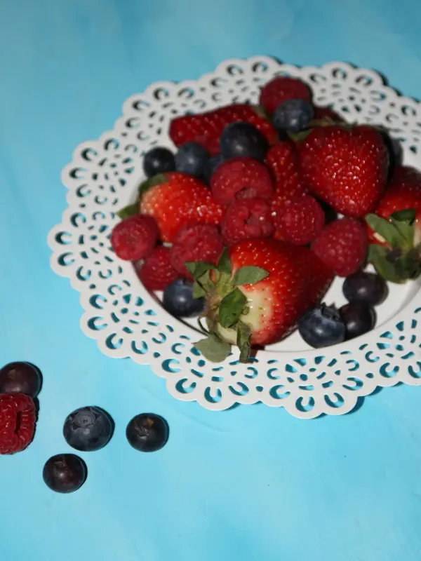 Eine dekorative, weiße Untertasse voller frischer Erdbeeren, Himbeeren und Blaubeeren auf einem hellblauen Tisch.