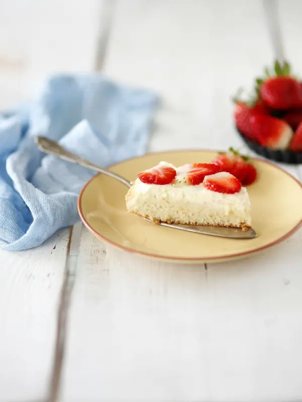 Ein Stück Kuchen mit Erdbeeren liegt auf einem Teller, während ein silberner Löffel daneben liegt und eine blaue Decke im Hintergrund zu sehen ist.