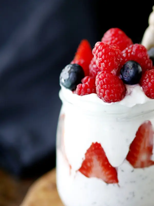Ein Glas mit cremigem, weißem Joghurt, garniert mit frischen Erdbeeren, Himbeeren und Heidelbeeren, steht vor einem dunklen Hintergrund.