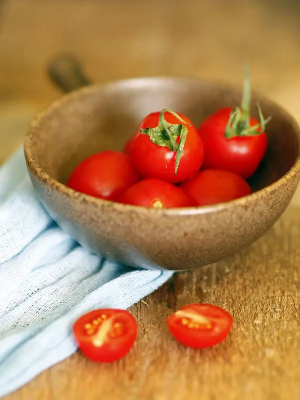Rote Tomaten liegen in einer braunen Schüssel auf einem Holzuntergrund, während einige halbierte Tomaten daneben liegen.