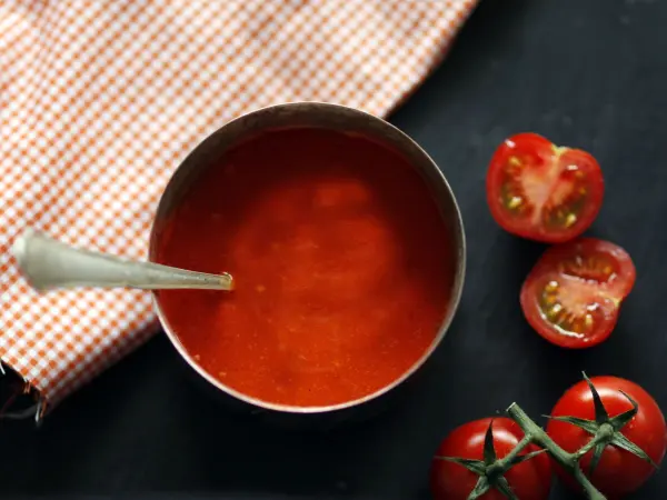Eine Schüssel mit Tomatensauce steht auf einem dunklen Tisch, umgeben von frischen, halbierten Tomaten und einem karierten Tuch.