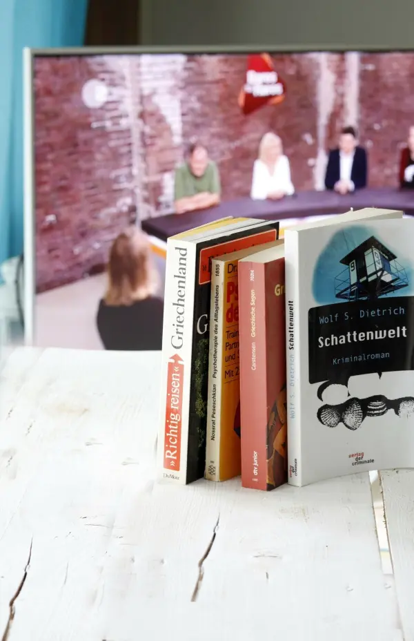 Fünf Bücher stehen ordentlich auf einem Tisch, während im Hintergrund eine Paneldiskussion auf einem Fernseher zu sehen ist.