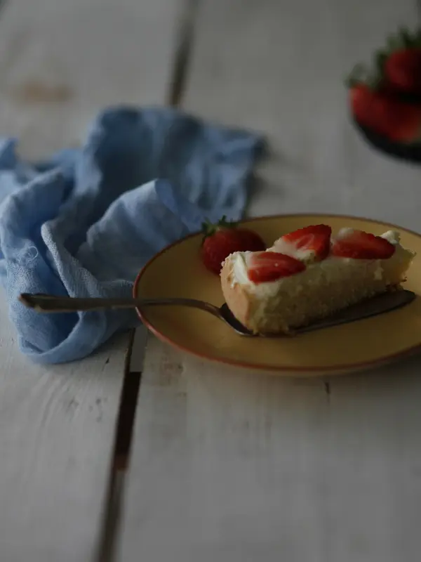 Ein Stück Käsekuchen liegt auf einem goldfarbenen Teller, garniert mit Erdbeeren, auf einem rustikalen weißen Tisch. Ein blaues Tuch ist darum drapiert.