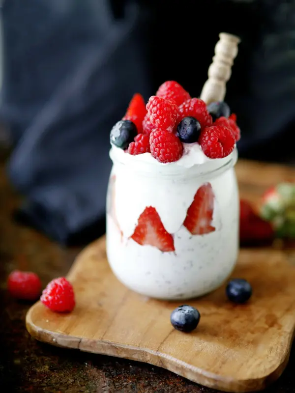 Ein Glas mit cremigem Dessert und frischen Beeren (Himbeeren, Blaubeeren) steht auf einem Holzbrett in einem dunklen Hintergrund.