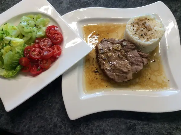 Ein saftiger Schweinsbraten liegt auf einem Teller mit Soße, begleitet von Reis und einem frischen Salat.