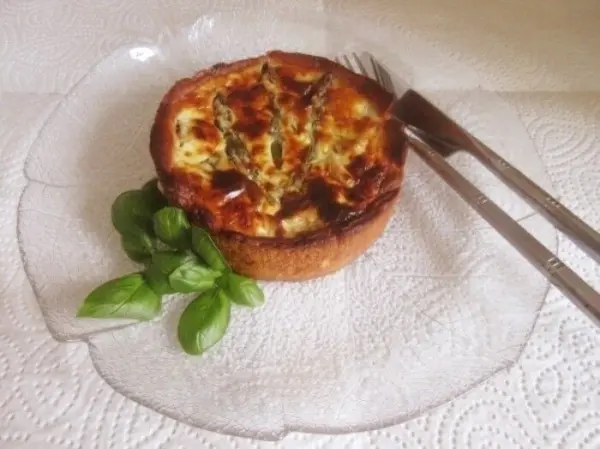 Eine runde Spargel-Quiche auf einem eleganten Glas-Teller, garniert mit frischem Basilikum und Besteck daneben.