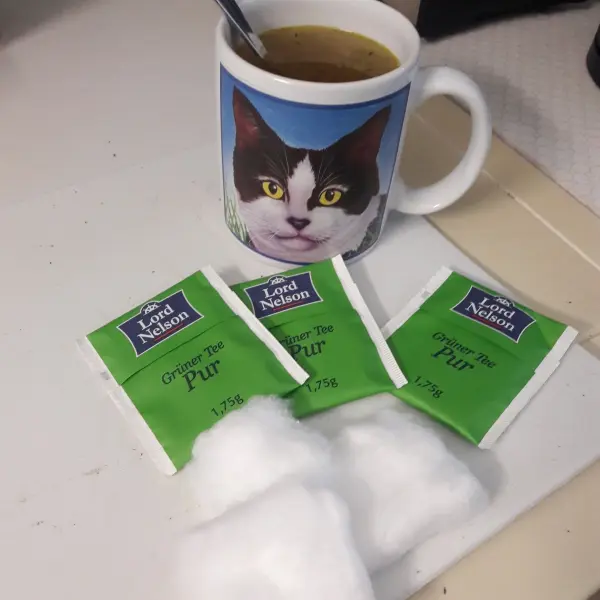 Ein Kaffeebecher mit einem Katzengesicht steht neben grünen Teebeuteln, die für das Wimpernwachstum verwendet werden.