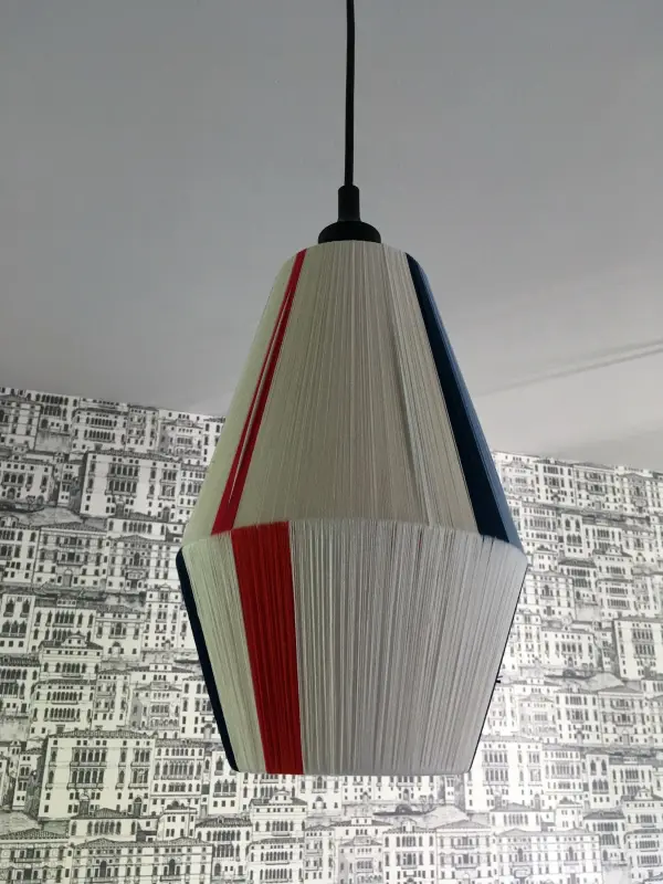 Ein hängender Lampenschirm, mit rotem, blauem und weißen Garn bespannt, schwebt vor einer grauen, grafischen Tapete.