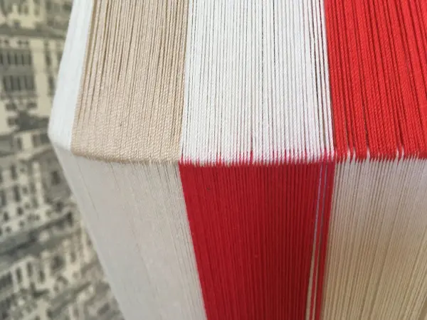 Ein Lampenschirm ist mit verschiedenen Garnen in Rot, Weiß und Beige bespannt und steht vor einem neutralen Hintergrund.