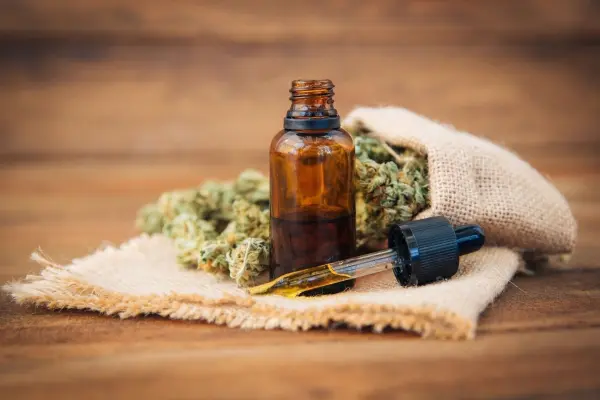 Ein braunes Fläschchen mit Tropfer steht auf einer stofflichen Unterlage, umgeben von Cannabisblüten, vermittelt Harmonie.