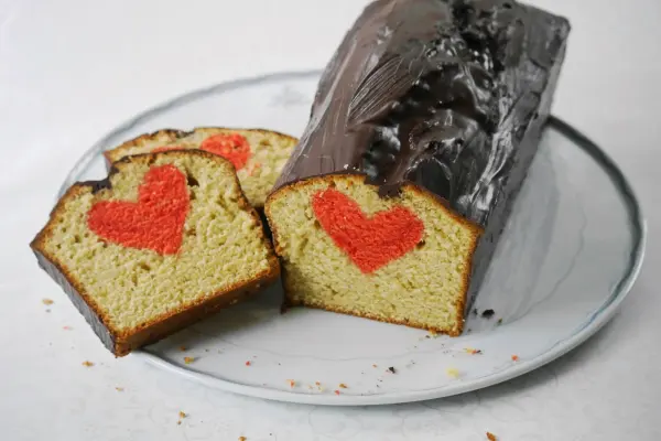 Ein Rührkuchen mit Herz, dekoriert mit roten Herzformen, auf einem weißen Teller, wird frisch angeschnitten.