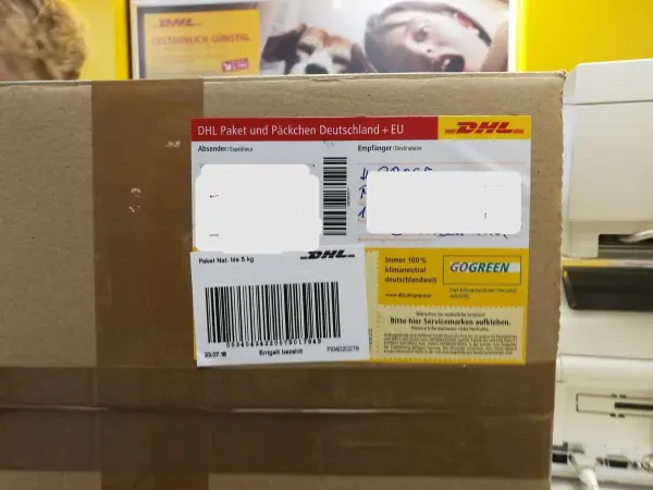 Ein braunes Paket mit DHL-Etikett liegt auf einem Tisch in einem Postbüro, umgeben von gelben Wänden und Druckern.