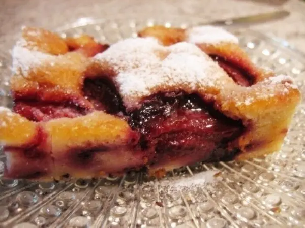 Ein Stück Obstkuchen vom Blech liegt auf einem gläsernen Teller, bestäubt mit Puderzucker und umgeben von Küchenutensilien.