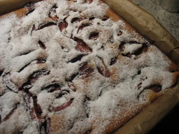 Ein Obstkuchen vom Blech liegt in einer Backform, mit einer zuckrigen Oberfläche und fruchtigen Stückchen sichtbar.
