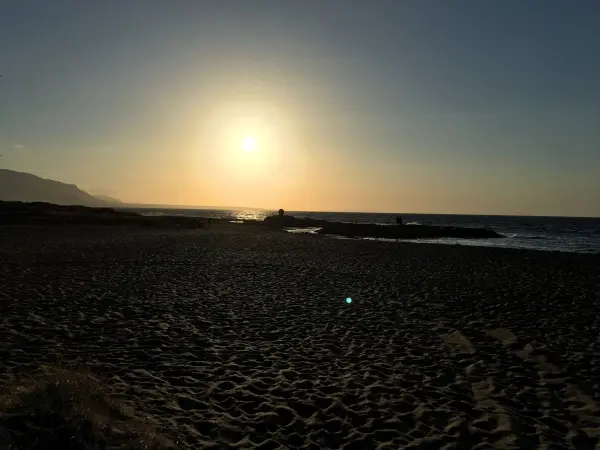 Die Sonne geht über dem Meer unter, während der Sandstrand in ruhiger Abendstimmung liegt. Meer und Küste sind sichtbar.