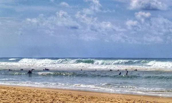 Surfer gleiten über die Wellen am Meer, während strahlender Himmel und sanfte Wolken eine entspannte Küstenatmosphäre schaffen.