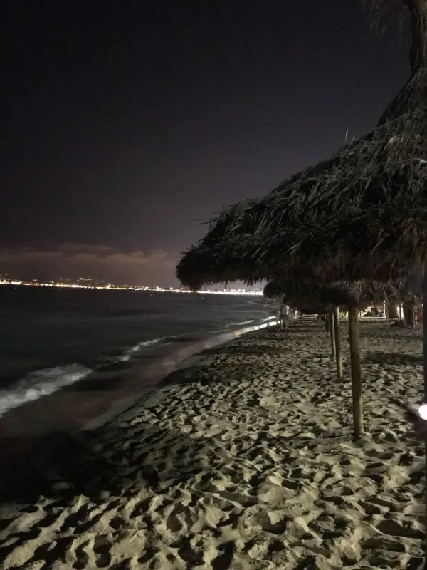 Am Strand stehen Strohdächer, während die Wellen sanft an den Sand schlagen und die Stadtlichter in der Ferne leuchten.