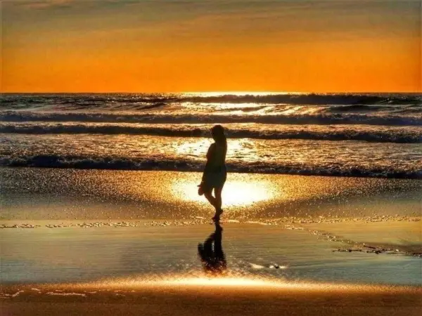 Eine Person spaziert am Strand, während die Sonne über dem Meer untergeht und den Himmel in orangefarbenes Licht taucht.