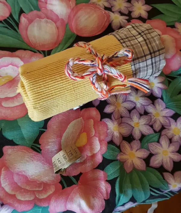 Ein gelbes Stoffetui für Nadelspiele ist mit einem karierten oberen Teil zusammengebunden auf einem bunten Blumenmuster platziert.